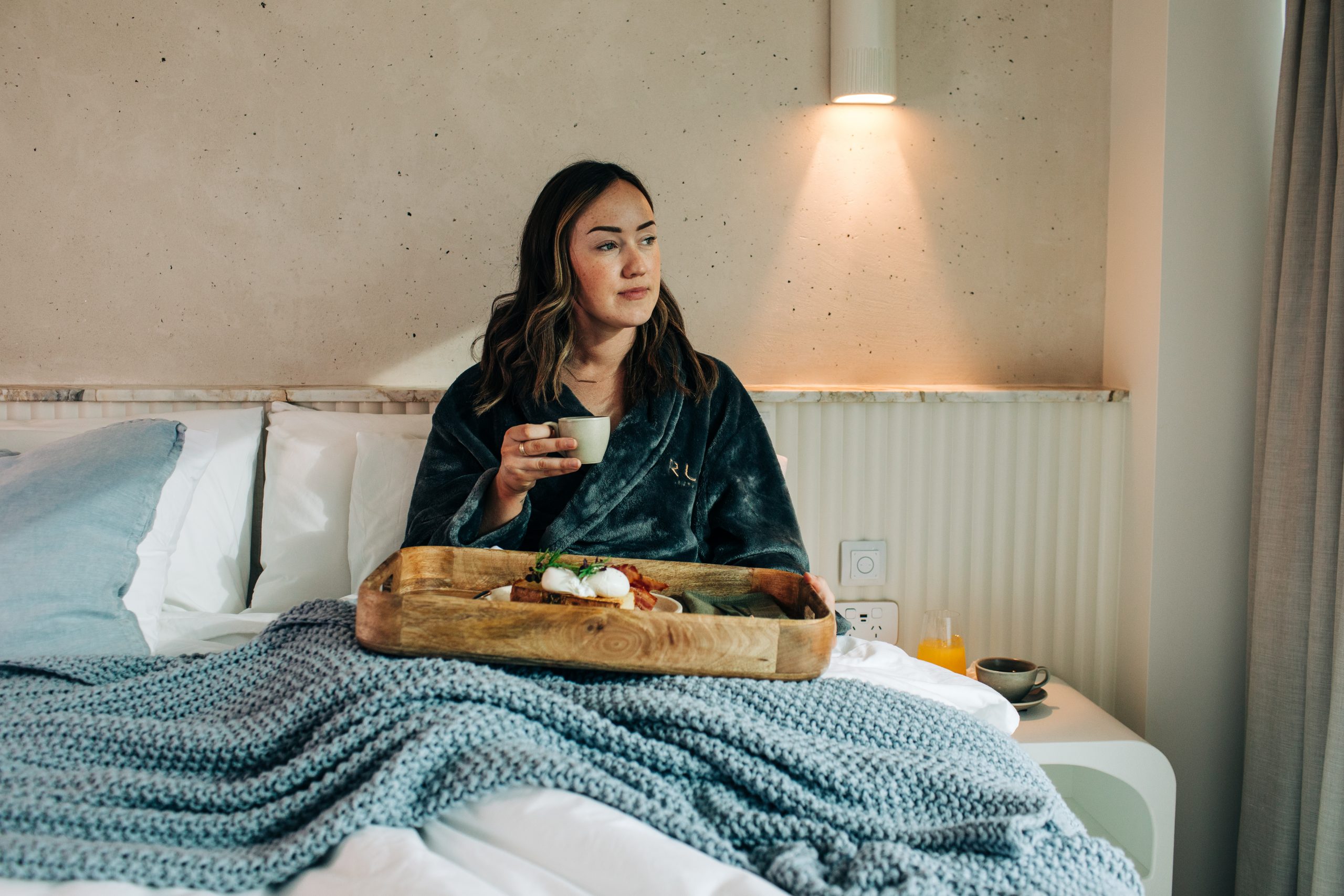 Woman sitting cozy in bed at Rumi on Louth private island retreat, enjoying a tray with a cup of tea while gazing out the window at the coastal view